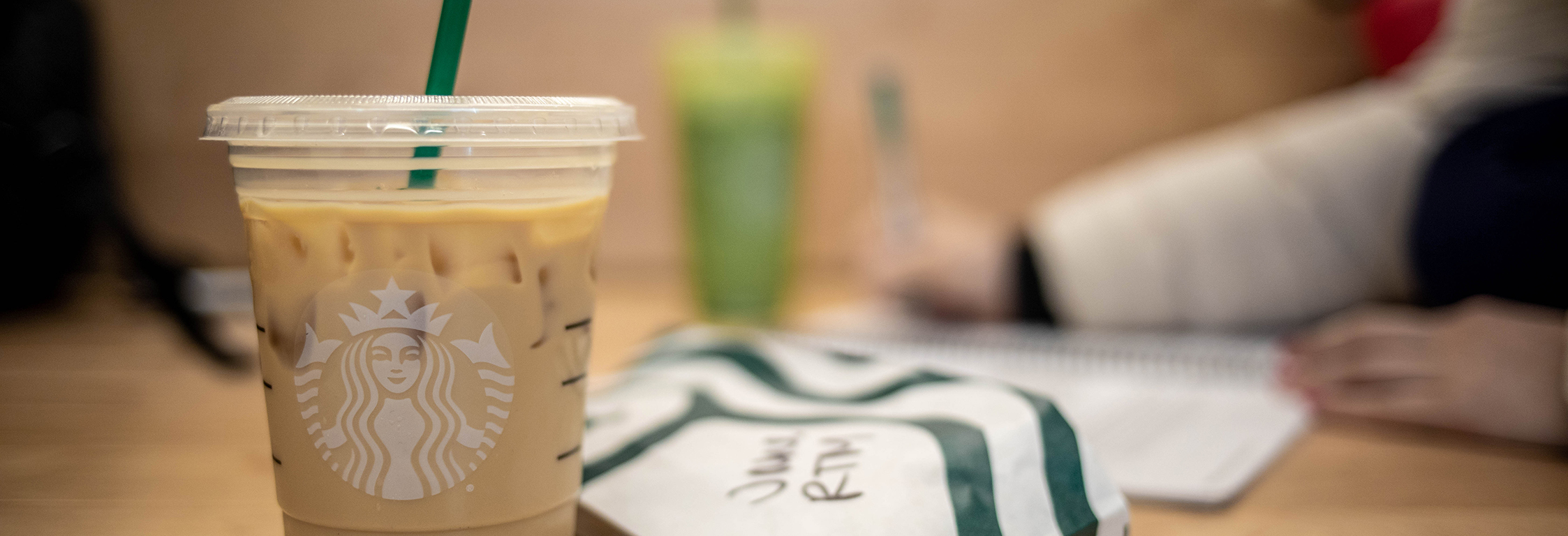 Iced Starbucks drink and wrapped food item sitting on a table. A person writes in the background.