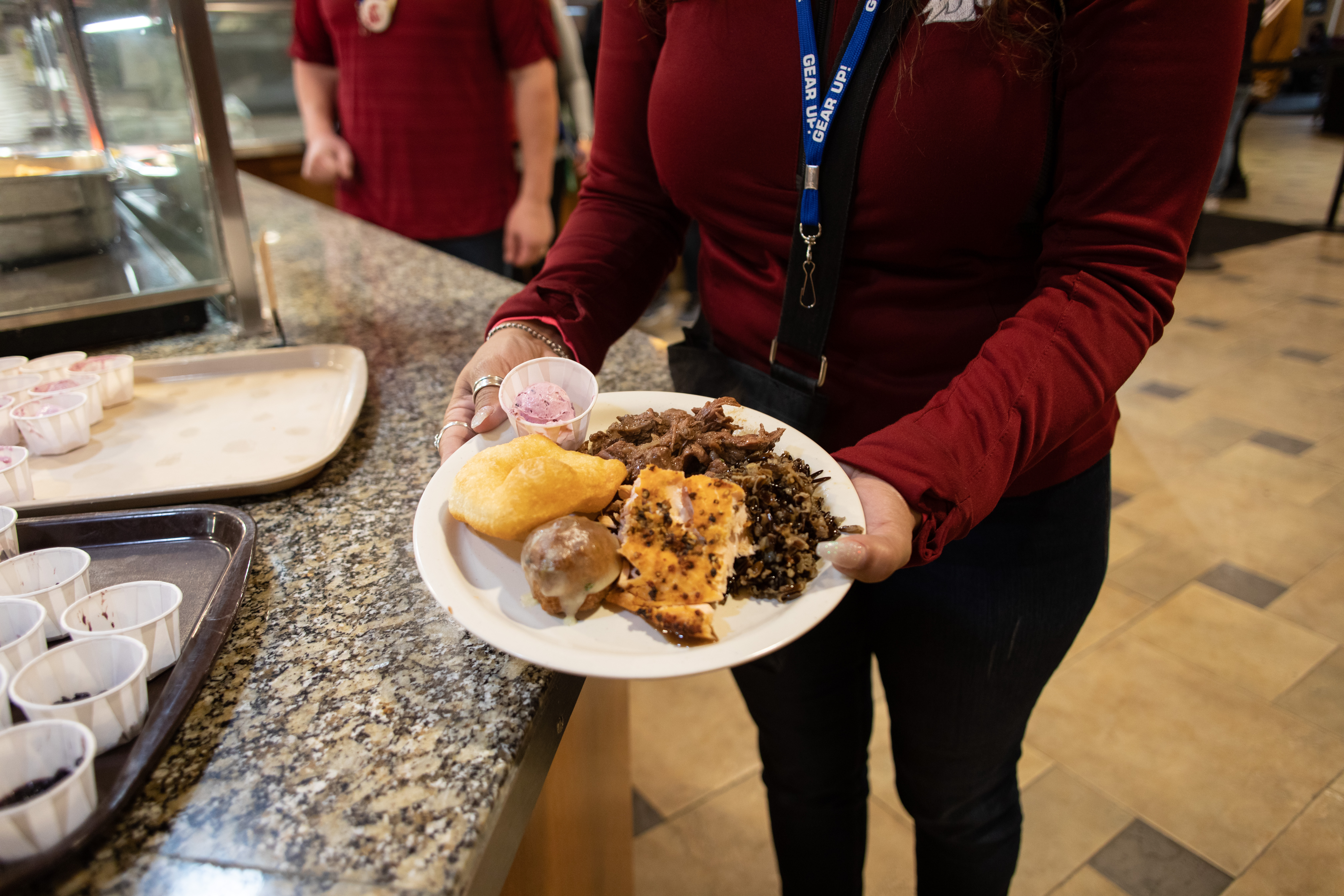 Shoulder down view of a person holding a plate of food in front of their belly button. She is wearing a red long sleeve shirt and black slacks. Her plate is round and white and filled with a yellow biscuit, some mixed green vegetables, a disposable paper sauce cup, a fried doughnut, and a meat dish.
