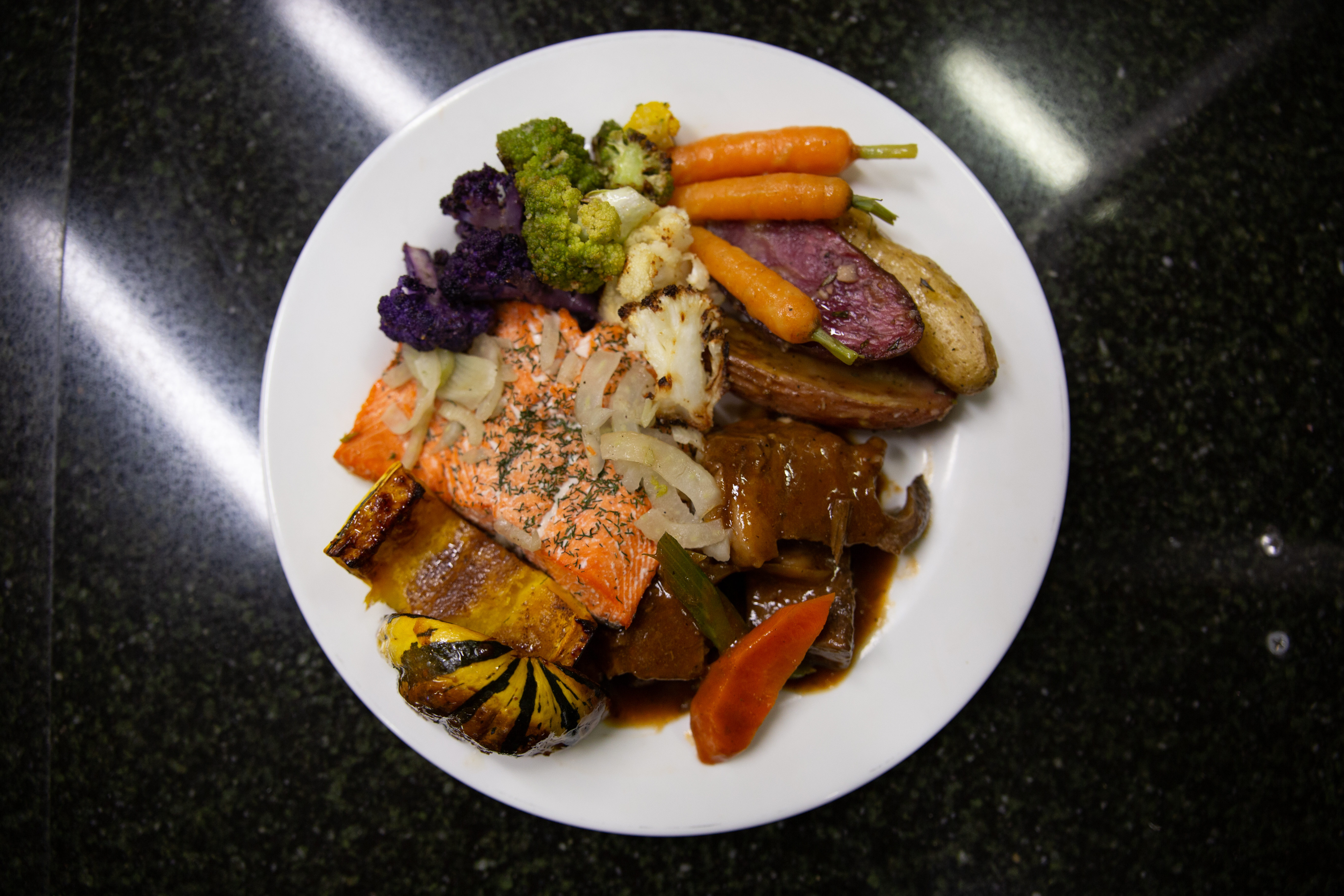 An overhead shot of a round white place on a large black countertop surface. On the plate is a very colorful assortment of fall vegetables: whole orange carrots, green broccoli, purple cauliflower, roasted purple fingerling potato, a roasted acorn squash cut in half to reveal orange flesh. The veggies are surrounding a pinkish fillet of salmon topped with a white sauce and green herbs.